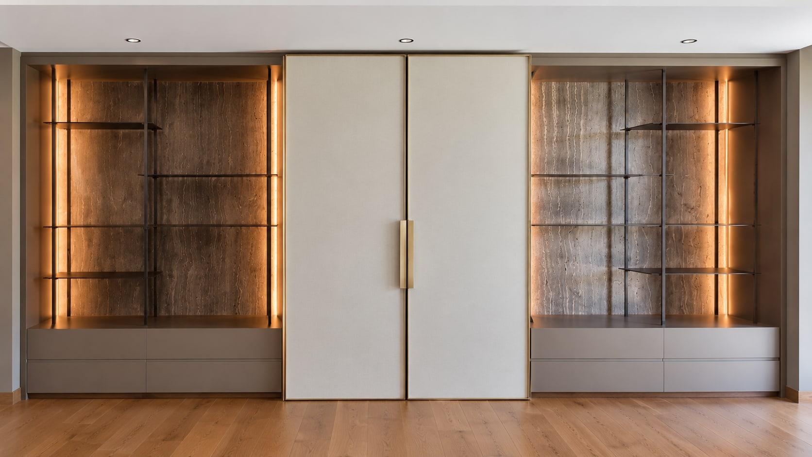 The bar wall closed: two ivory doors in brass frames between lit travertine shelving niches