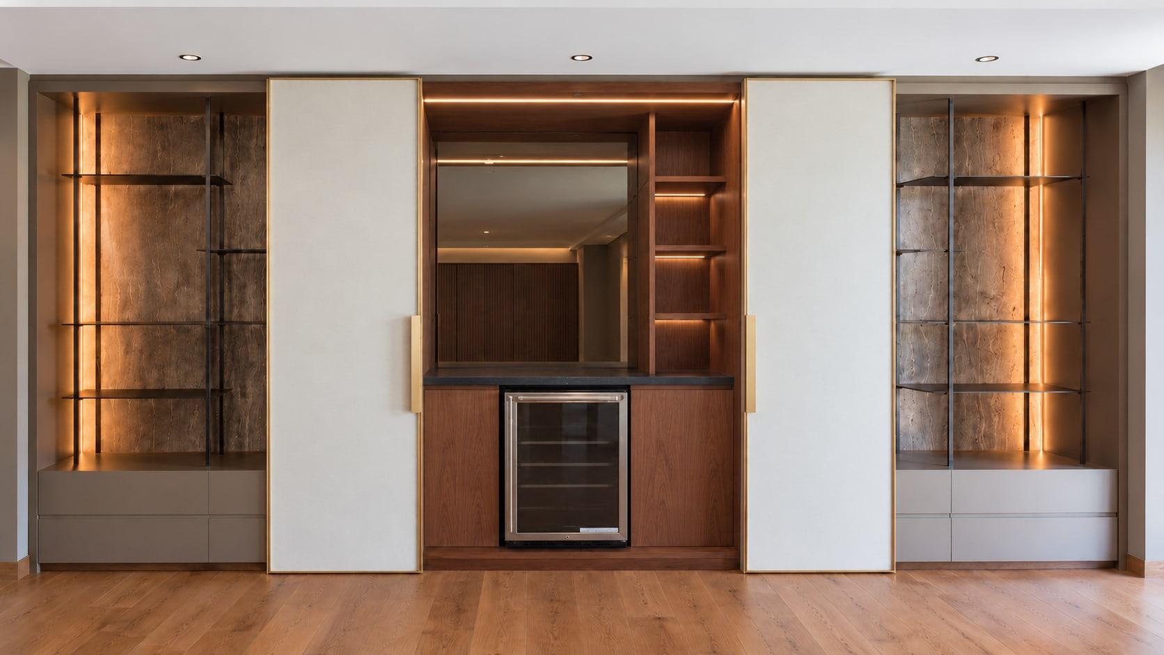 A walnut bar between two ivory leaves slid open: a mirror, lit shelves and a wine fridge