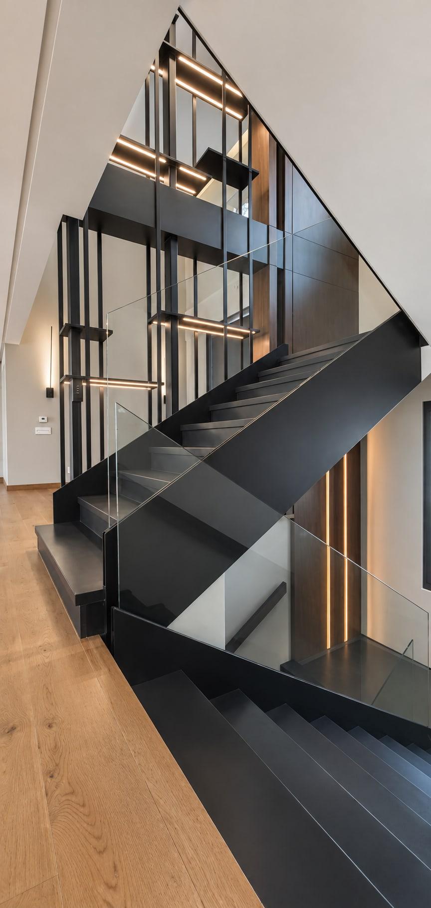 Black steel stair turning down inside the steel screen, with lit shelves and a glass balustrade, seen from above