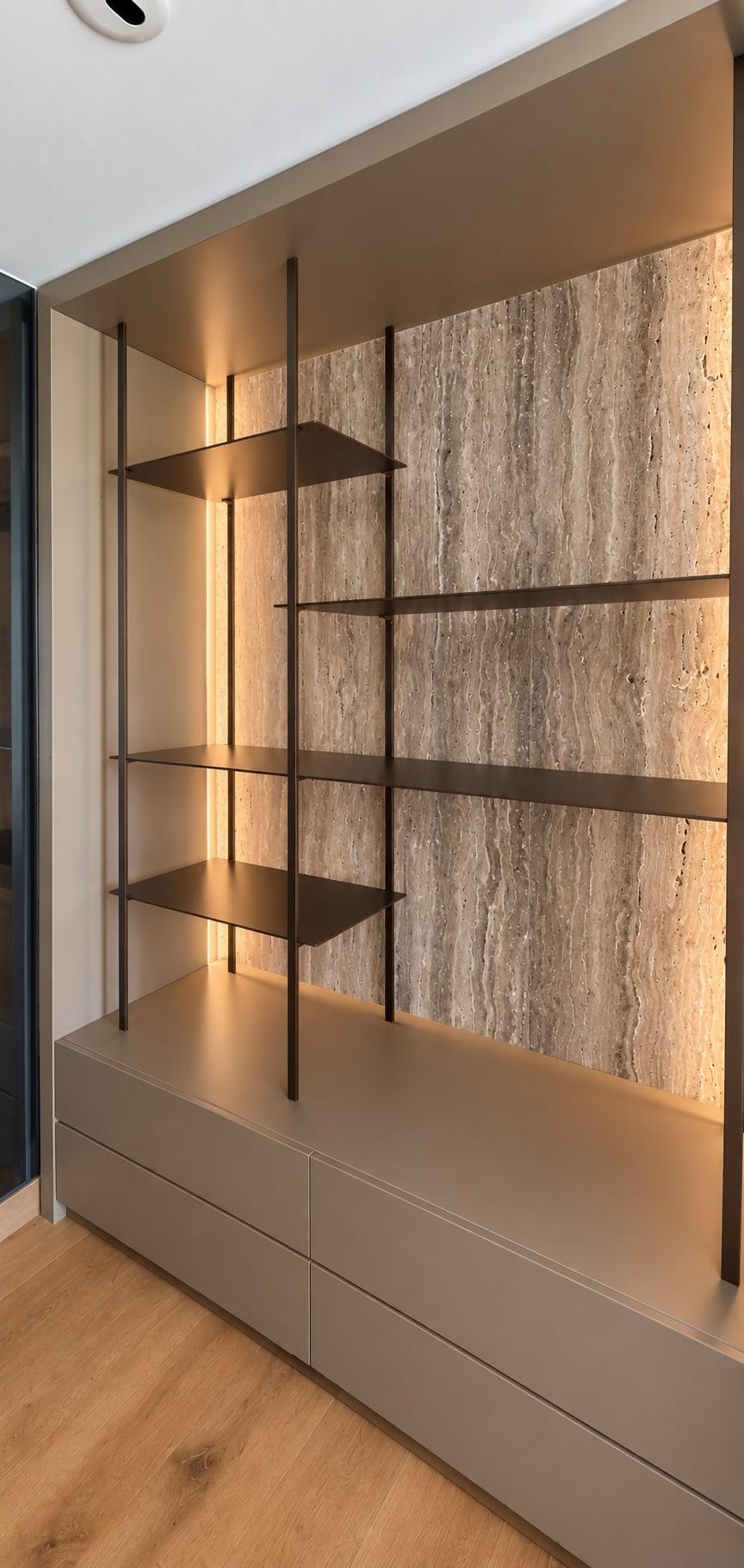 Shelving niche backed in travertine with dark metal shelves and side lighting, over a taupe lacquer drawer base