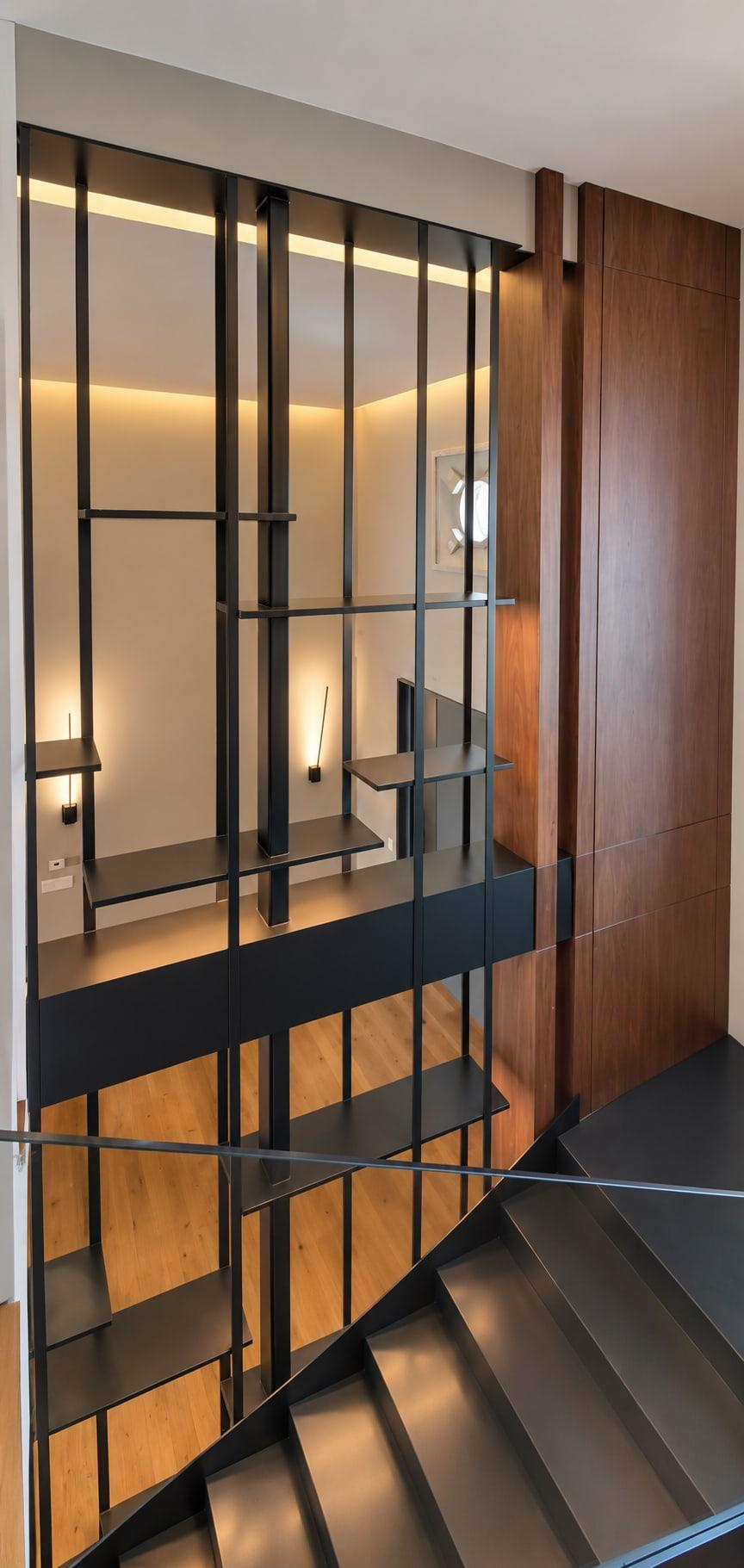 Blackened steel stair screen with lit shelves beside walnut panelling
