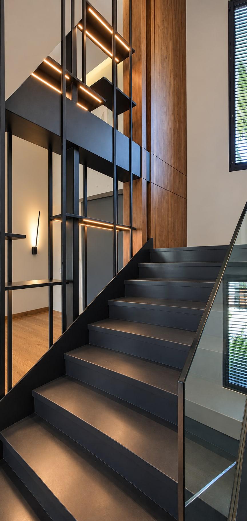 The same stair finished: blackened steel and walnut beside the flight, lit shelves and a glass balustrade