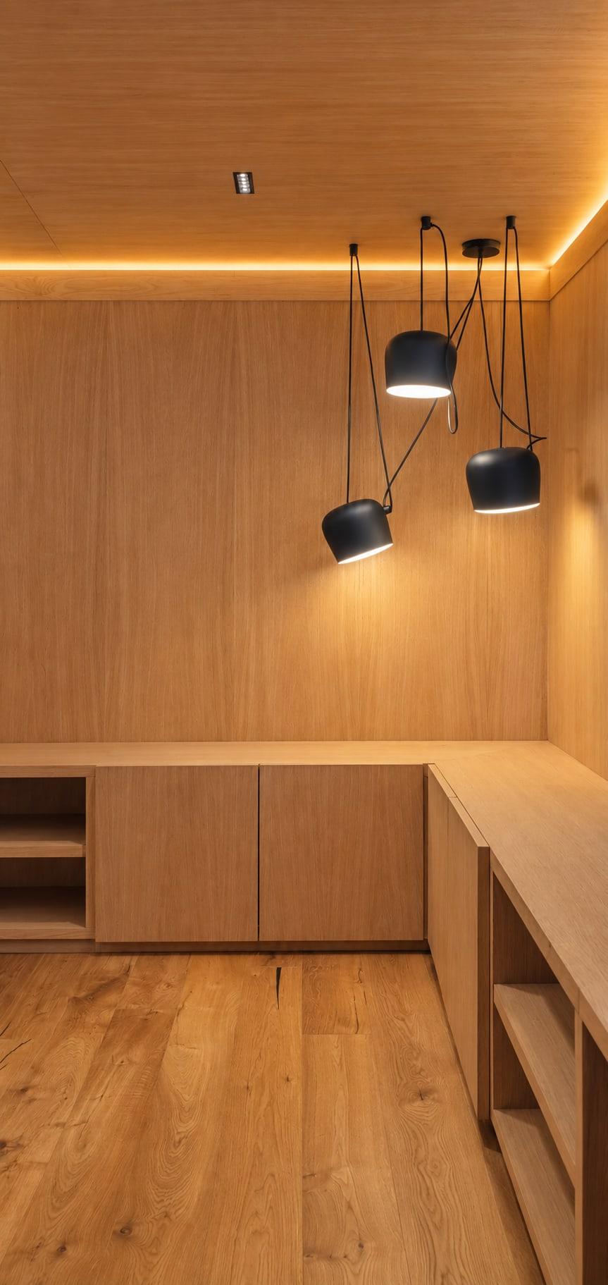 Oak-panelled study with built-in cabinets and an L-shaped desk run under three black pendants and a cove light