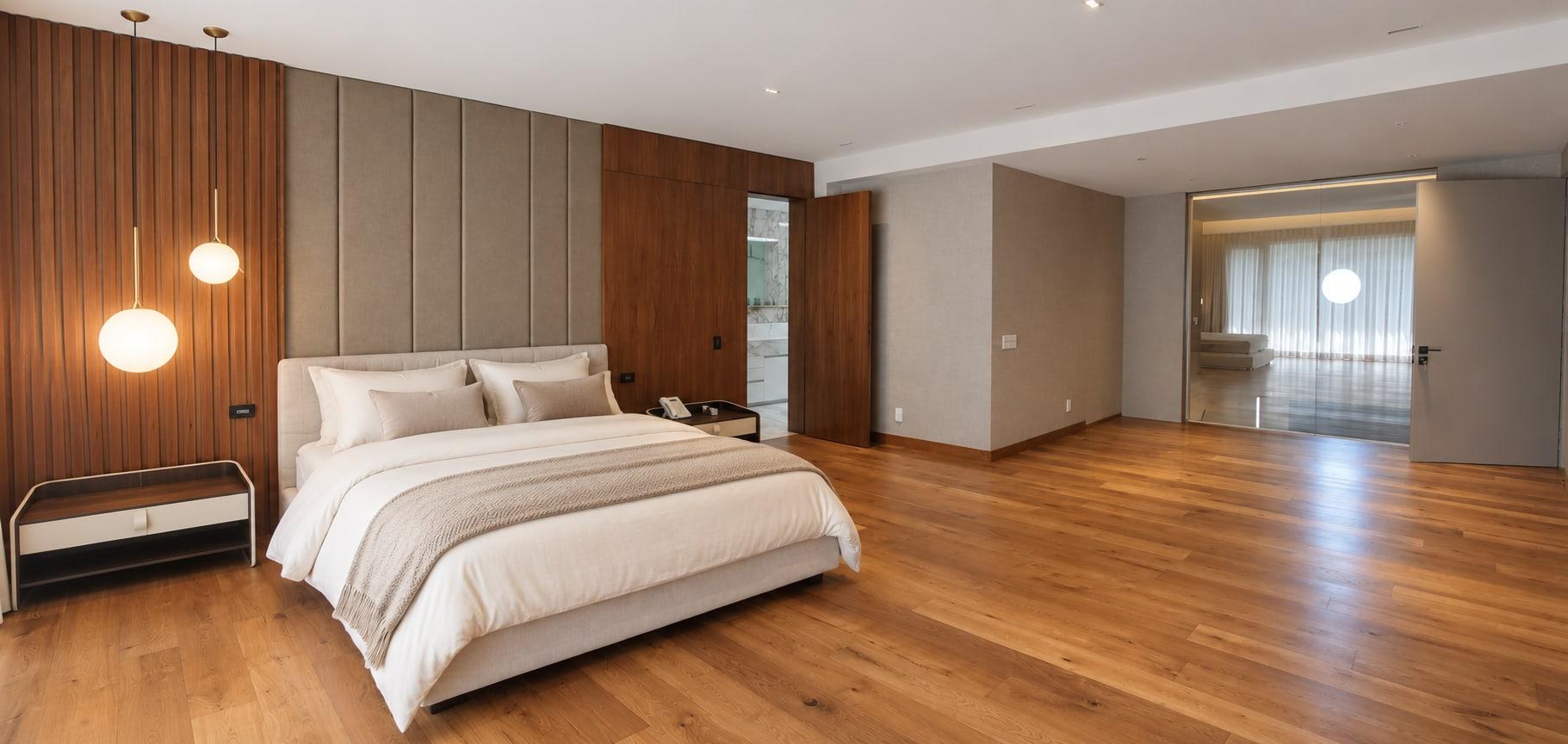 The finished primary suite: an upholstered headboard wall between walnut slats, a walnut door and glass into the next room