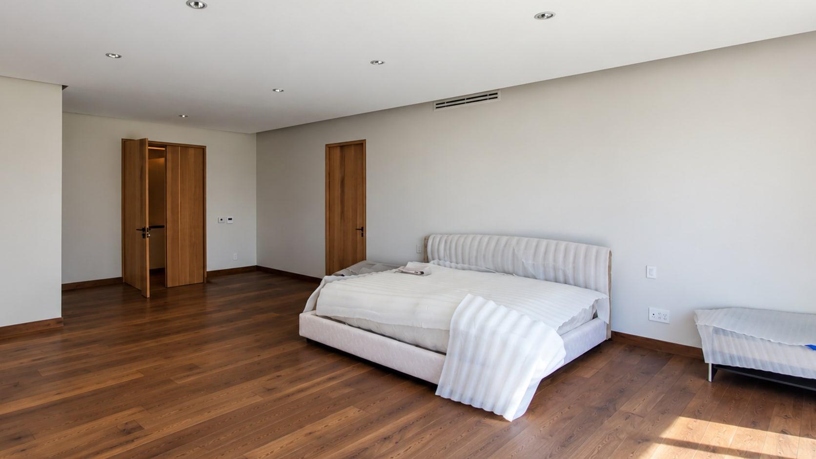 The primary suite as received: a bed in its packing against a plain wall under a ceiling grille, a dark wood floor and two oak doorways