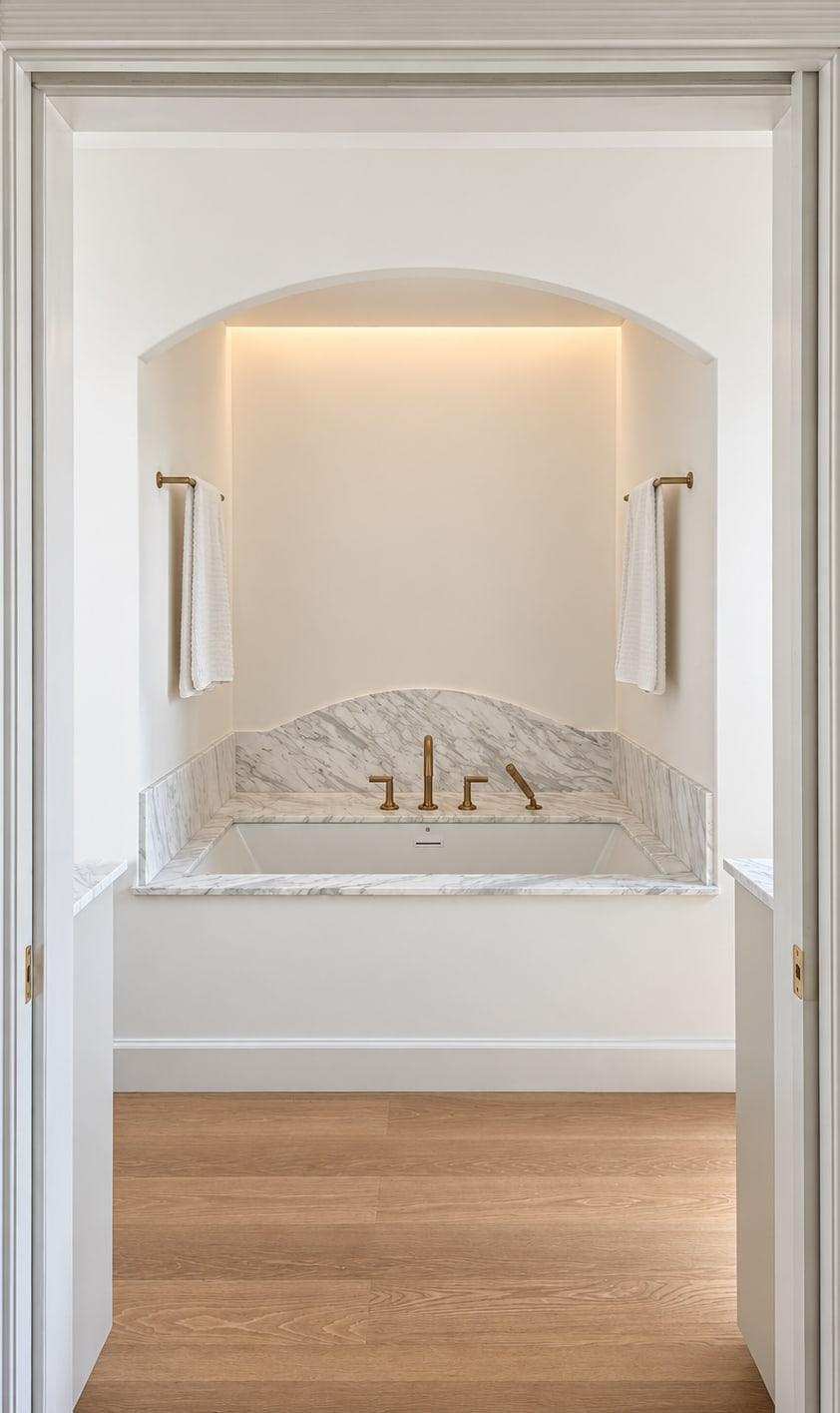 The same bath finished, through the same door: a tub set in white marble in a lit plaster alcove, with brass fittings and a wood floor
