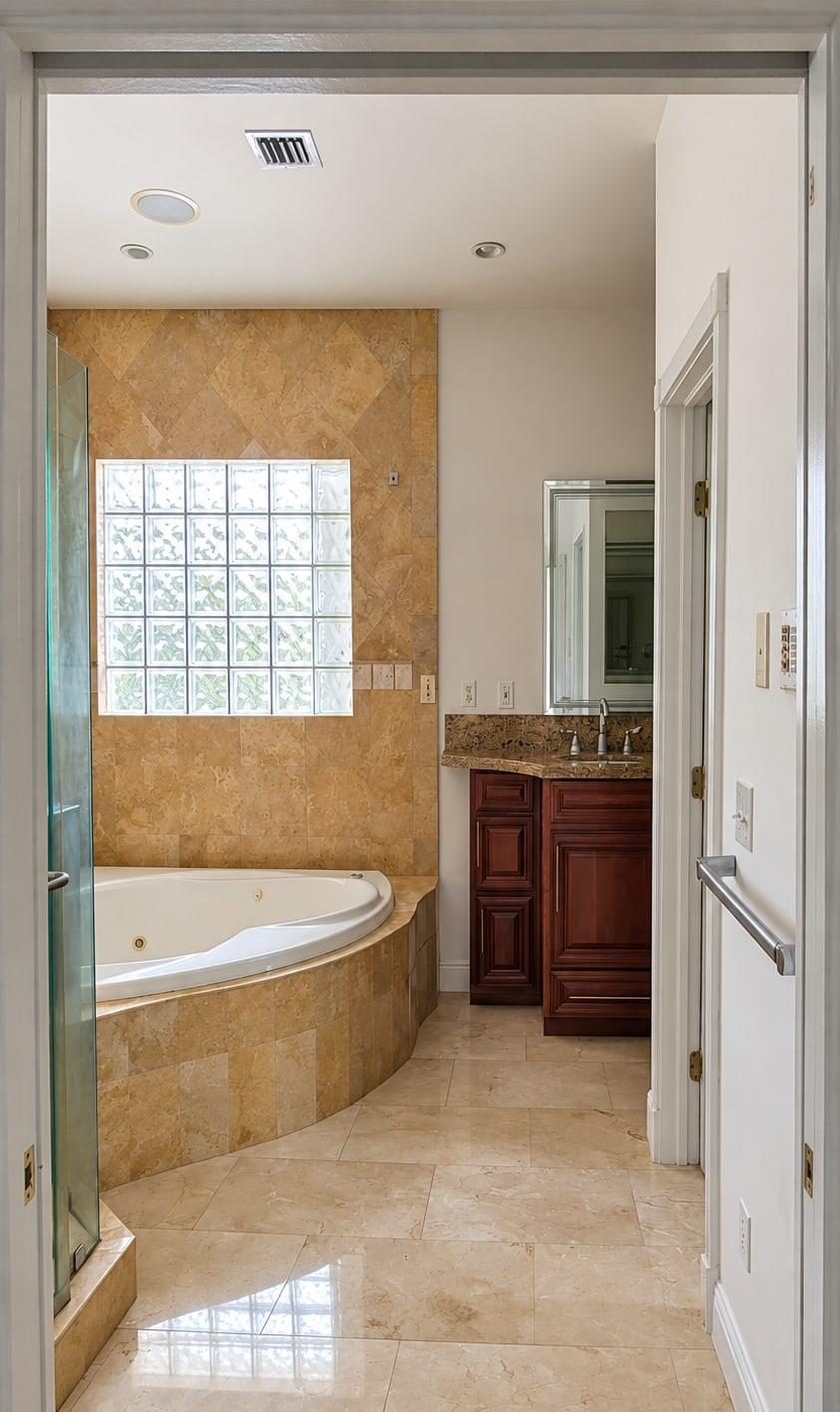 The bath as received, through the door: travertine walls, a corner jacuzzi under a glass-block window and a cherry vanity