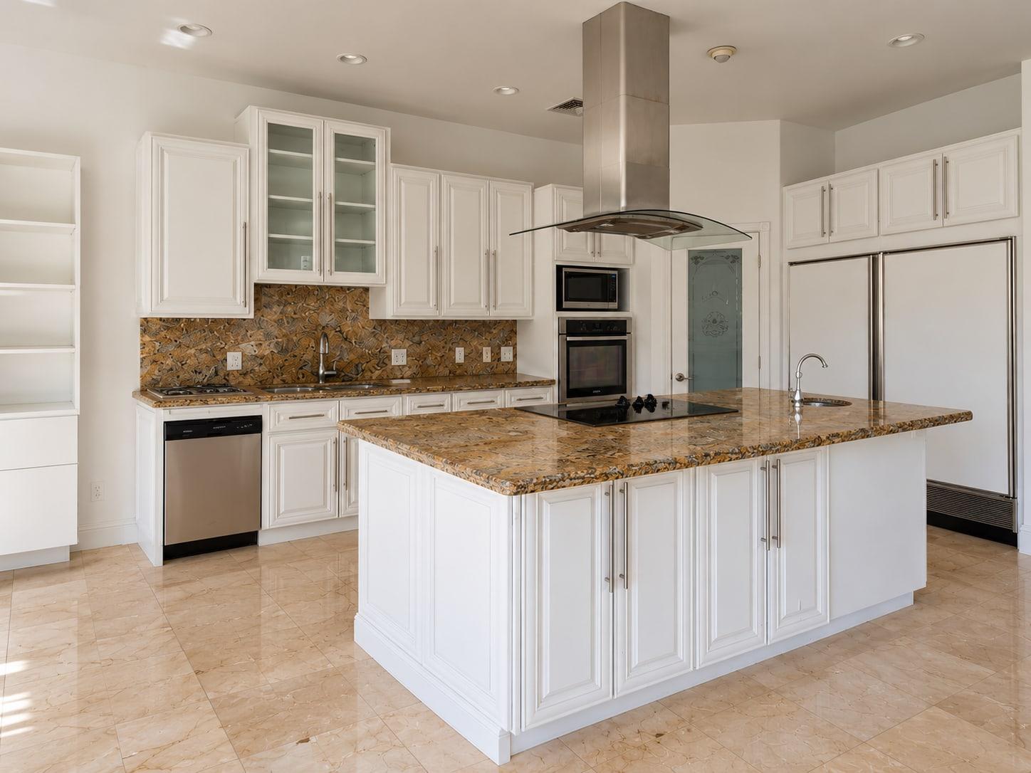 The kitchen as received: glass-fronted white cupboards over a stone mosaic splashback, stacked wall ovens and a steel hood over the granite island