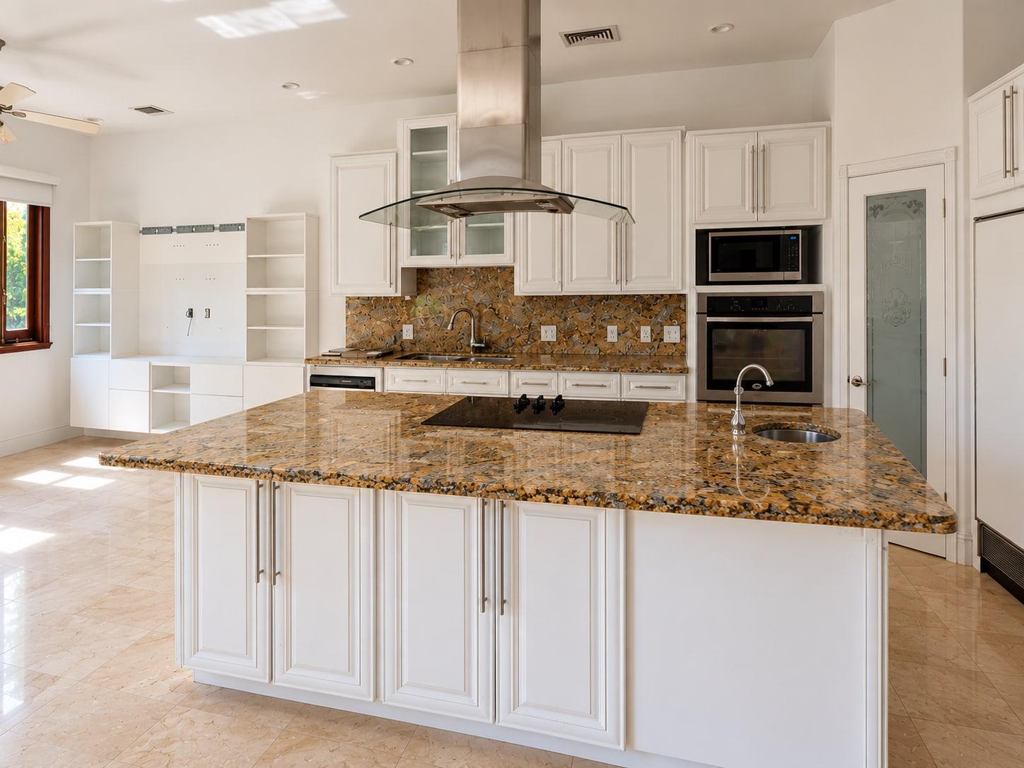 The island as received: white raised-panel cupboards under a brown granite slab, with a hob and a sink set into it and a steel hood above