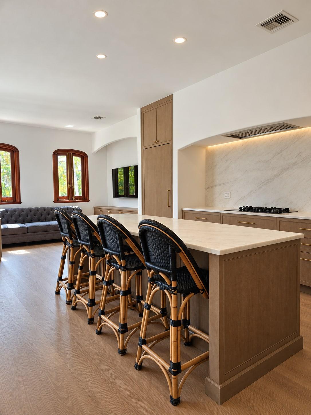 Oak kitchen island with a pale stone top and four black rattan stools, the living room and two arched windows beyond