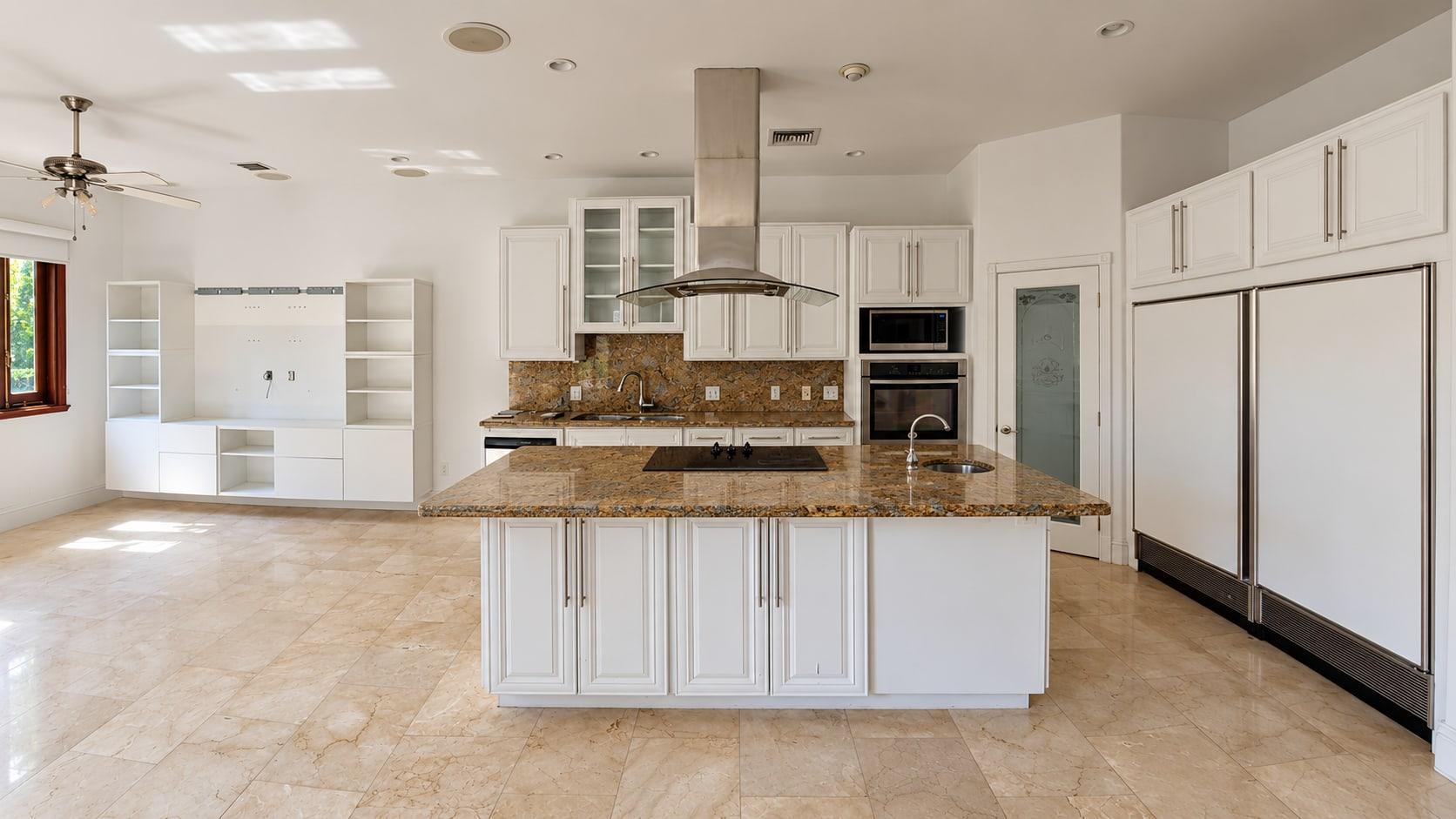 The kitchen as received: white raised-panel cupboards, a brown granite island under a steel hood and a marble tile floor