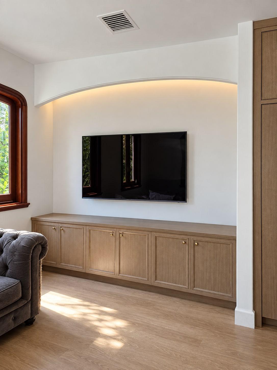 Low oak media cabinet under a cove-lit plaster arch, with a screen on the wall above and a grey sofa beside it