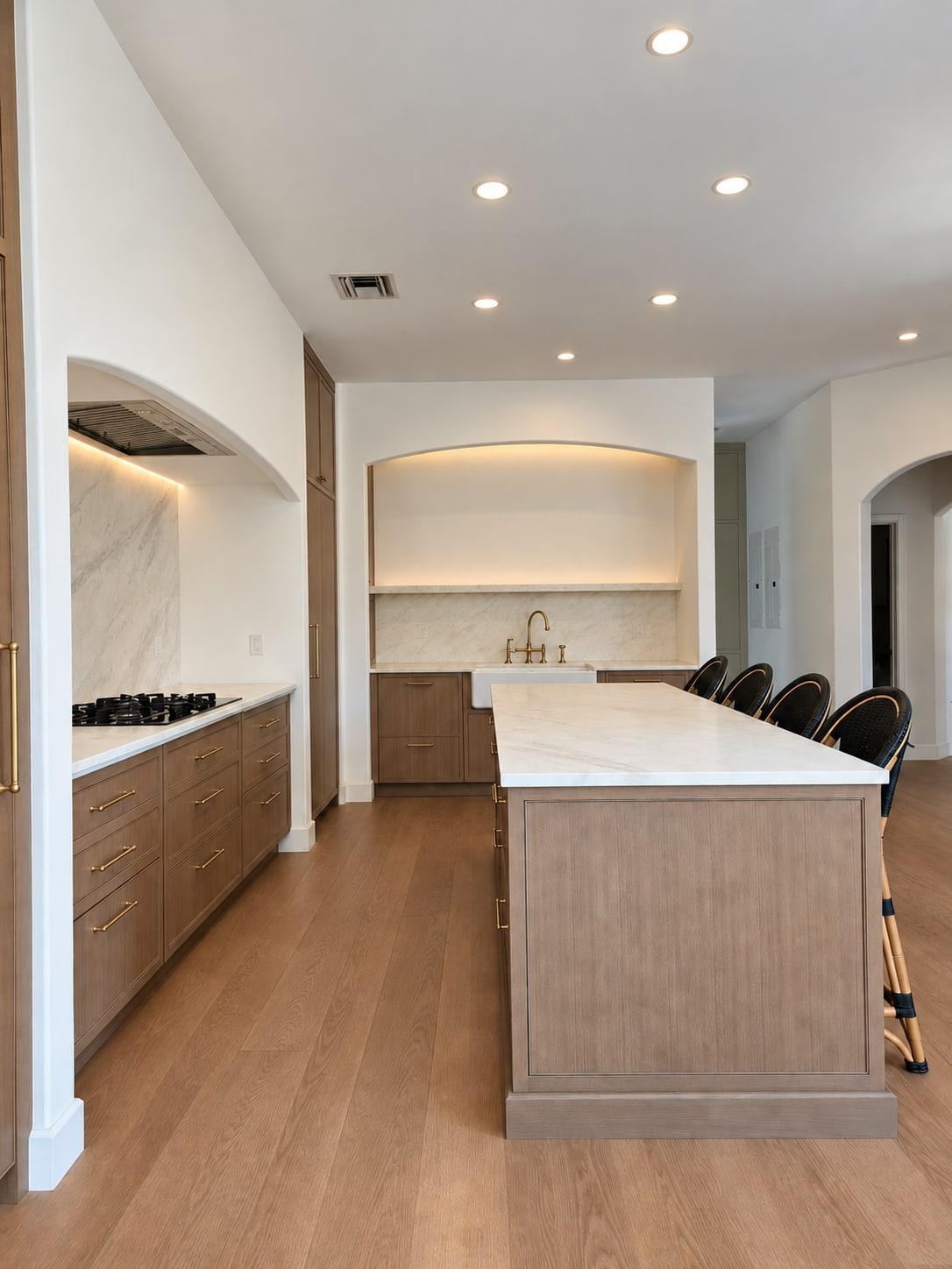Oak kitchen run with a gas cooktop under a lit plaster arch, the island in the middle and the sink alcove at the far end
