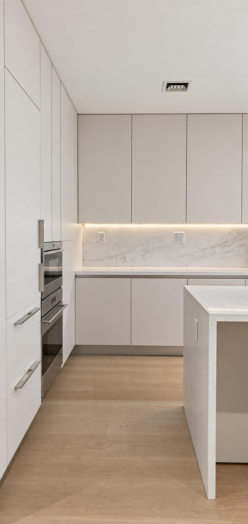 Tall run of matt lacquer units holding wall ovens beside a lit splashback