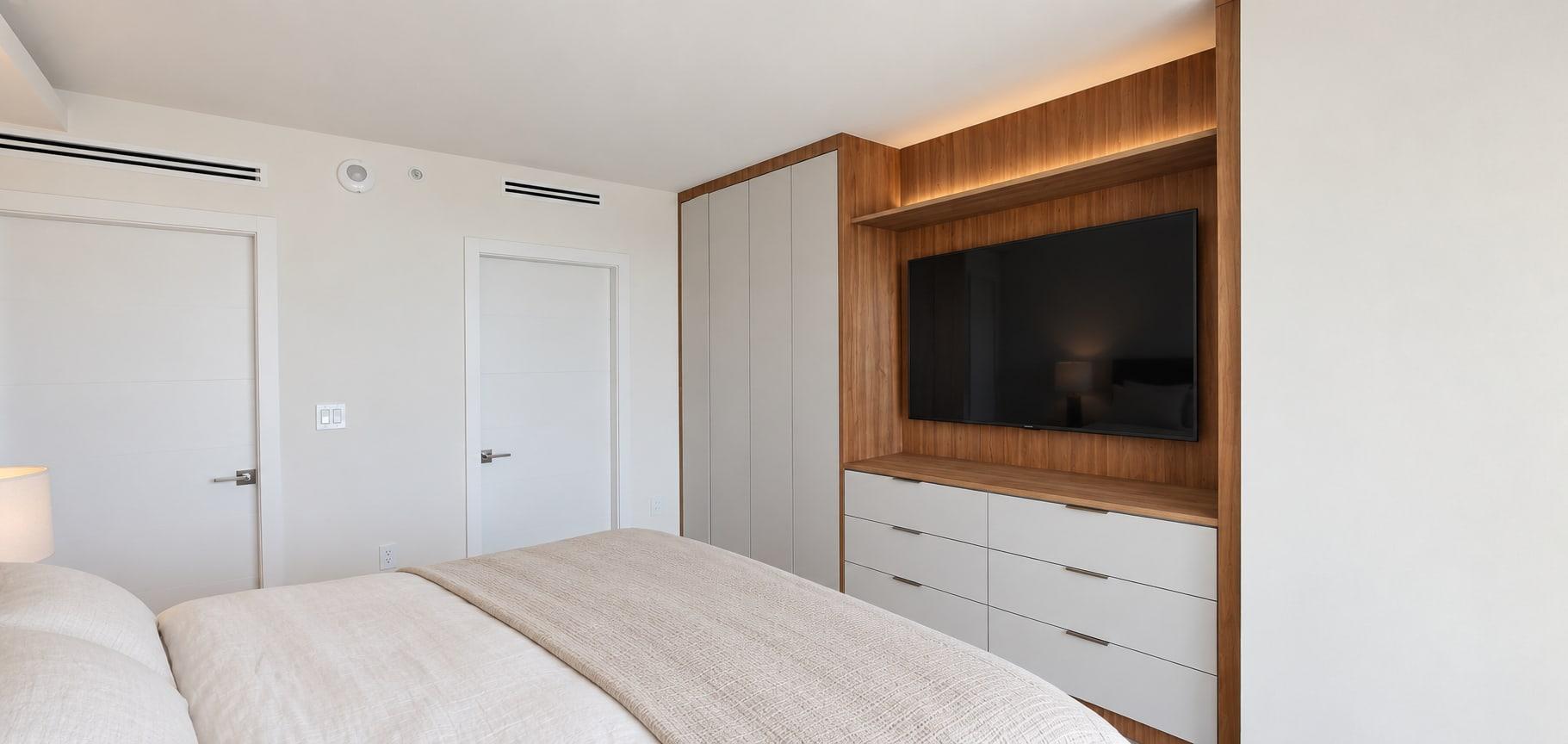 Bedroom with a walnut-lined niche holding the screen, lit along its head, over a run of white lacquer drawers beside a tall pale wardrobe