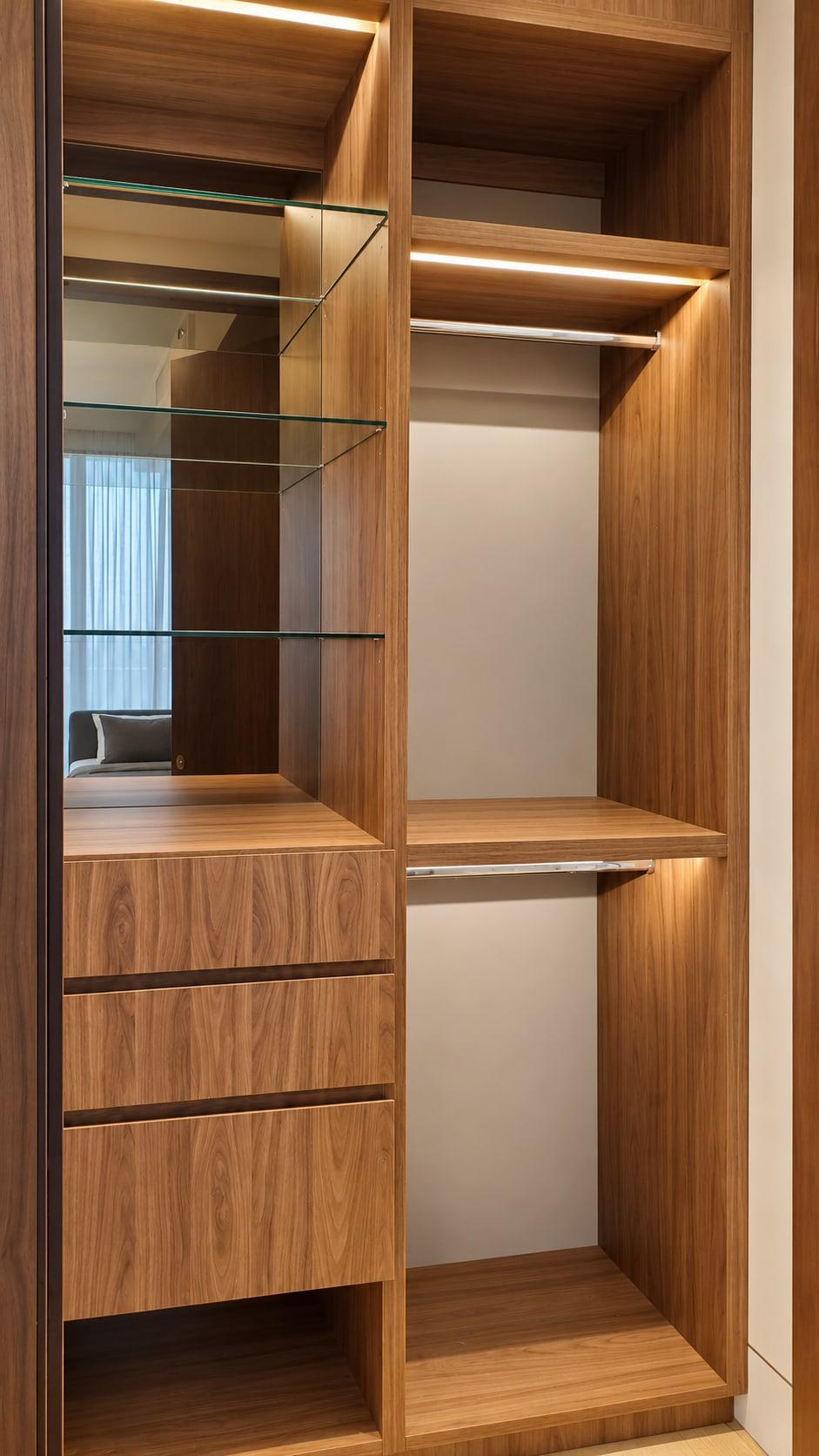 Walnut wardrobe bay with glass shelves in front of a bronze mirror, over walnut drawers, a lit hanging rail beside it