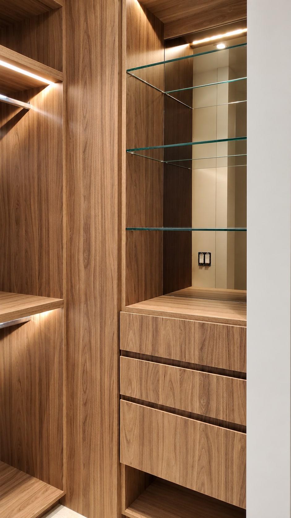 Walnut wardrobe bay with glass shelves over a stack of drawers, beside a hanging rail lit from the shelf above