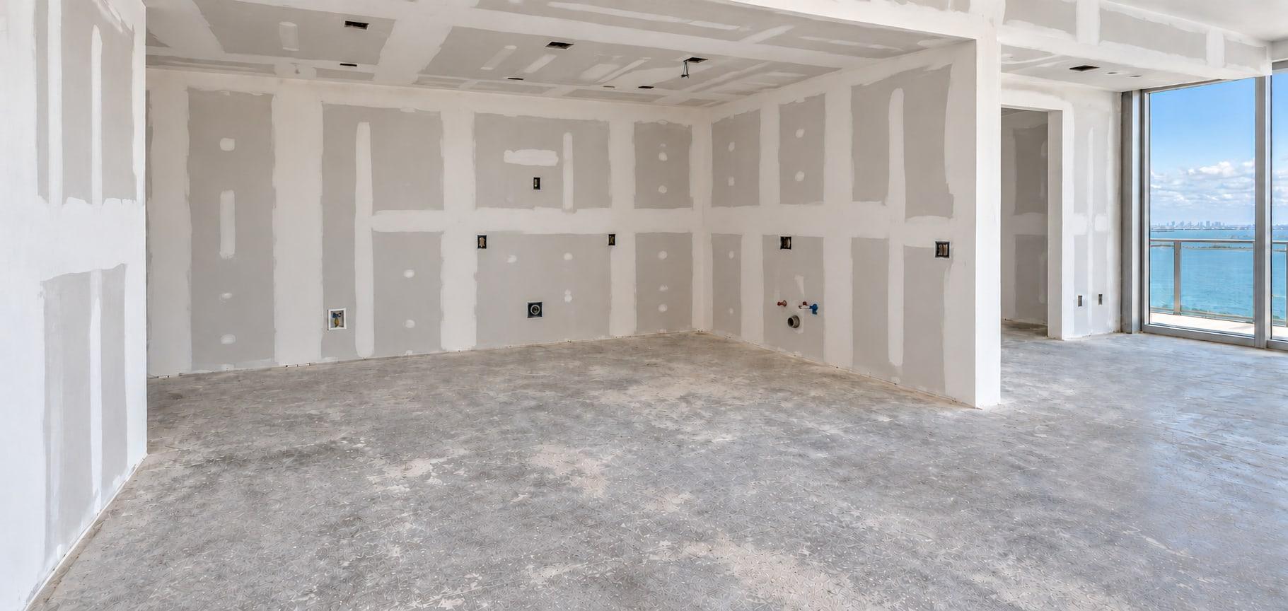The kitchen as received: taped drywall, a bare concrete slab, pipes and boxes in the far wall and the bay through the window on the right