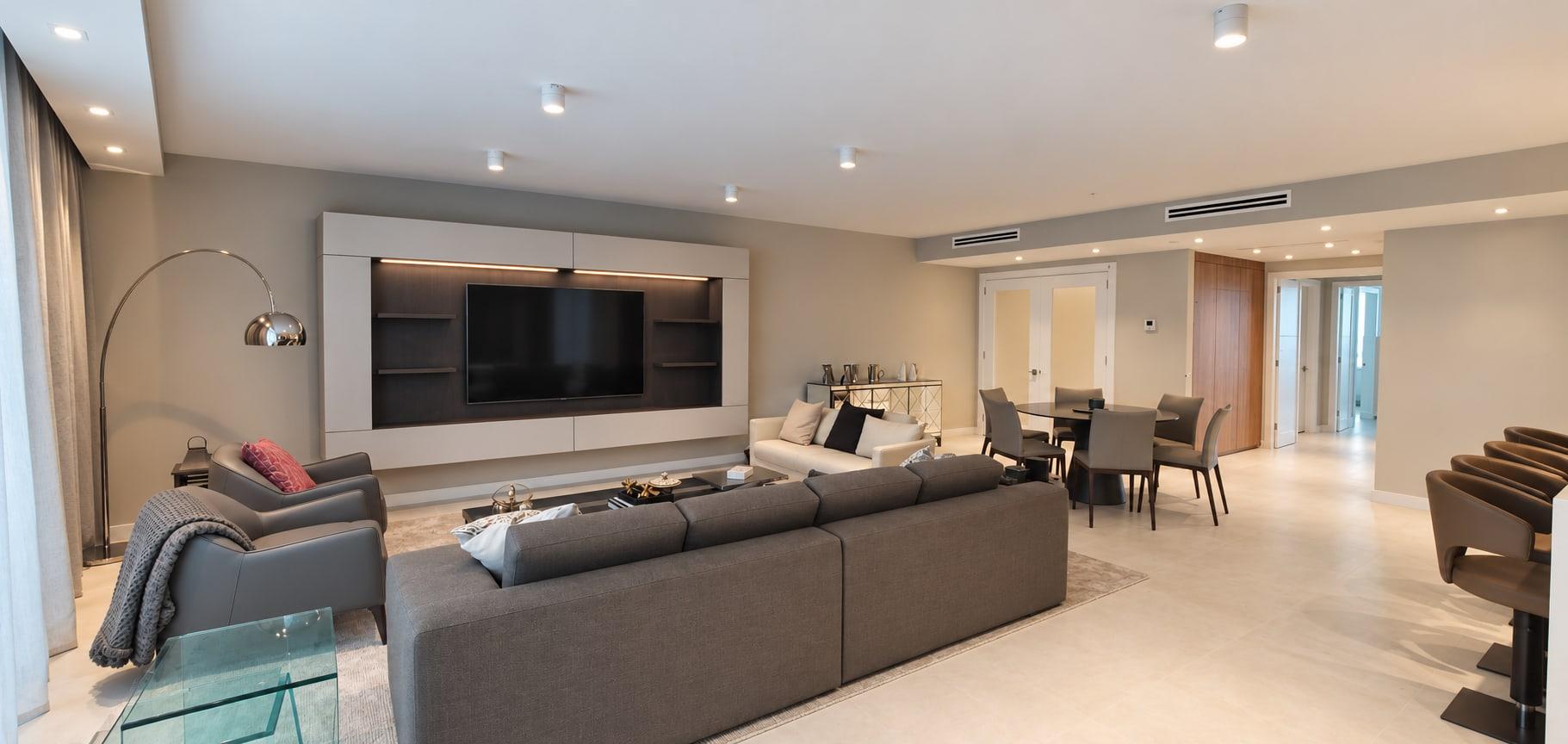The finished living room: the screen in a pale lacquer frame lit along its head, a grey sofa and armchairs, a dining table and glazed doors to the lift lobby