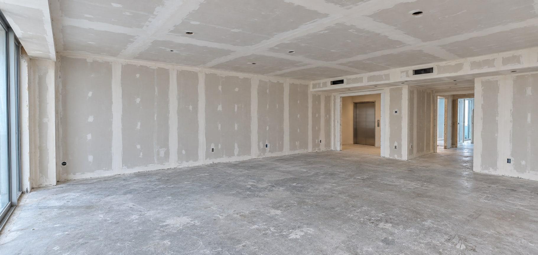 The living room as received: taped drywall on the walls and ceiling, a bare slab, windows down the left and the lift lobby open at the far end
