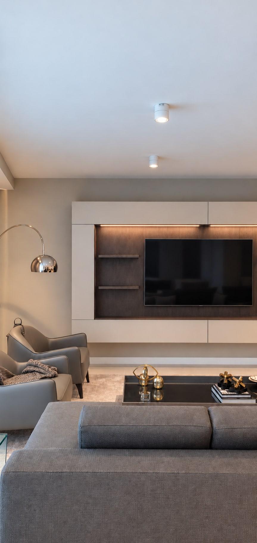 Media wall of pale lacquer framing a dark back with the screen and open shelves, lit along its head, over a grey sofa and armchairs