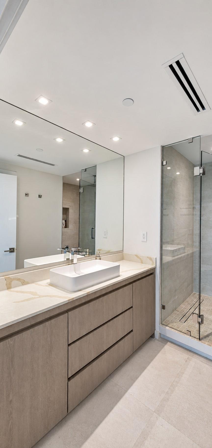 Vanity in taupe wood grain with a white marble top and a basin standing on it, under a full-width mirror, a glass shower beside it